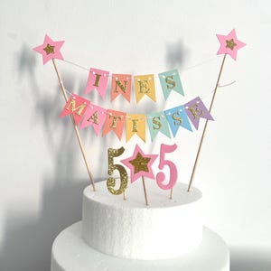 A cake topper featuring two wooden sticks supporting a multi-colored pennant banner that spells out INES MATISSE in gold glitter letters. Below the banner are two number 5s, one in gold glitter and one in pink, with a pink star in between.