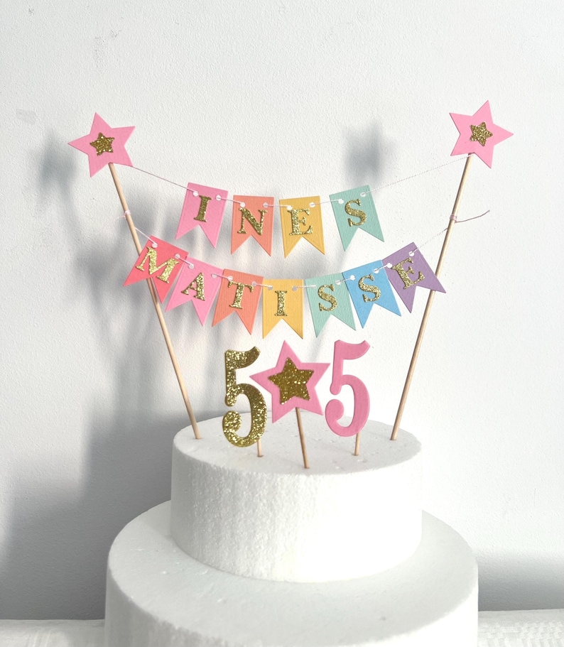 A cake topper featuring two wooden sticks supporting a multi-colored pennant banner that spells out INES MATISSE in gold glitter letters. Below the banner are two number 5s, one in gold glitter and one in pink, with a pink star in between.