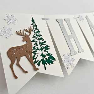 Pode incluir: Uma faixa branca com a palavra "Happy" em letras prateadas com glitter. A faixa é decorada com flocos de neve, uma árvore de pinho verde e um veado marrom.