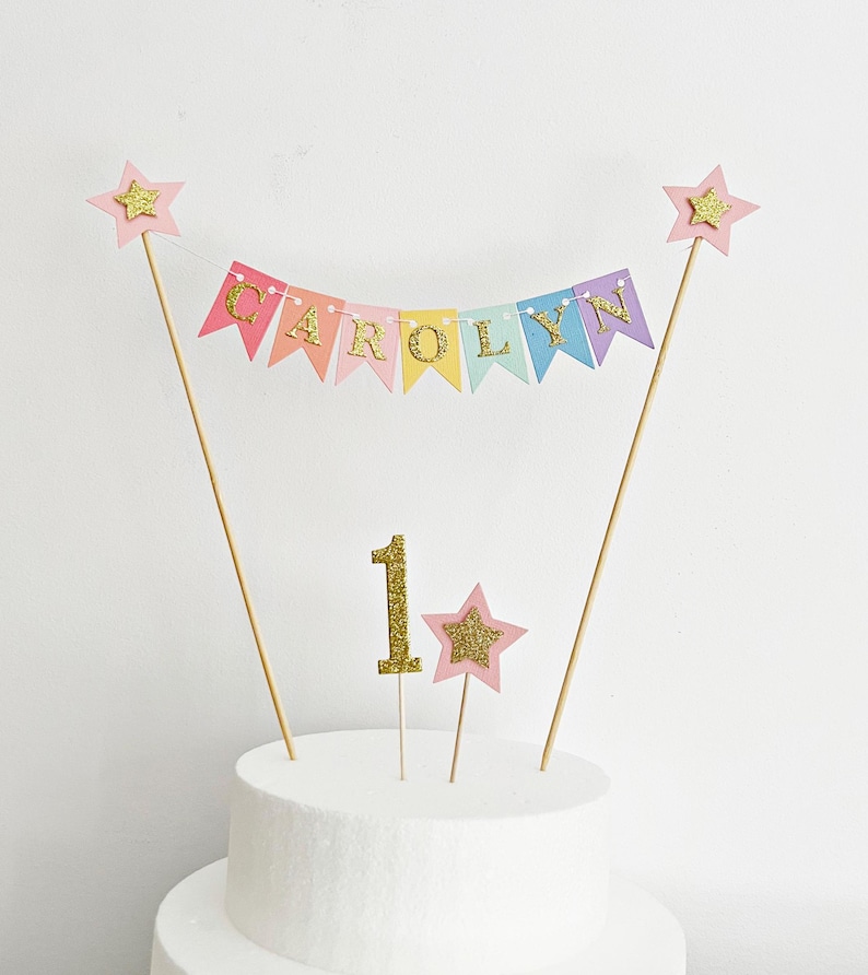 A cake topper set featuring a pastel bunting banner with the name CAROLYN in gold glitter letters, two wooden sticks topped with pink stars, a gold glitter number 1, and a small pink star topper, all displayed on a white cake.