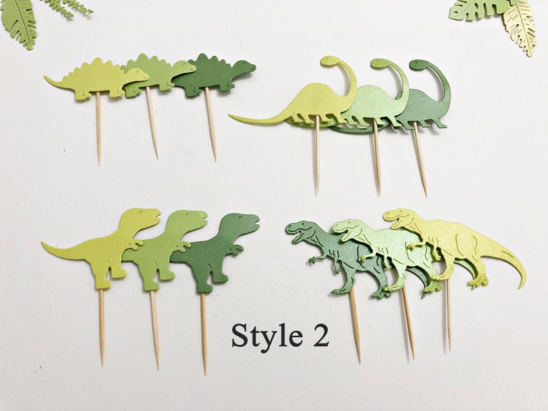 May include: A set of green dinosaur cupcake toppers. The toppers are made of cardstock and feature different dinosaur designs, including a stegosaurus, a brachiosaurus, and a tyrannosaurus rex. The toppers are perfect for adding a fun and festive touch to any dinosaur-themed party.