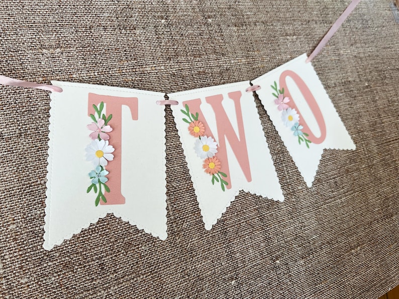 以下が含まれることがあります： ピンクと白の旗に、ピンクの文字で「TWO」と書かれており、小さな紙の花で飾られています。旗はピンクのリボンで結ばれています。