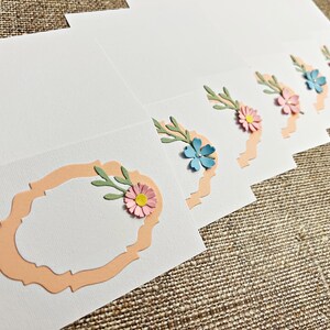 以下が含まれることがあります： 桃色の波状の枠と紙の花が付いた空白のカード6枚セット。花はピンク、ブルー、薄いピンクで、中心は黄色です。カードはメモやメッセージを書くのに最適です。
