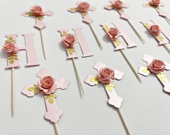 May include: Pink and gold cross and letter H cupcake toppers with a floral design. The cross and letter are made of paper and have a 3D effect. The flowers are pink and have a gold center. The toppers are perfect for a baptism or christening.