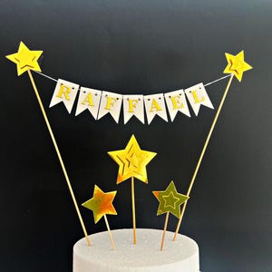 May include: A cake topper featuring a white bunting banner with the gold letters spelling RAFFAEL, suspended between two thin wooden sticks topped with gold stars. Three additional gold star picks of varying sizes are positioned below.