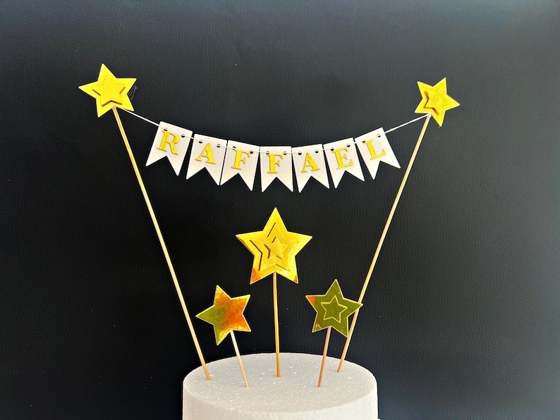 May include: A cake topper featuring a white bunting banner with the gold letters spelling RAFFAEL, suspended between two thin wooden sticks topped with gold stars. Three additional gold star picks of varying sizes are positioned below.