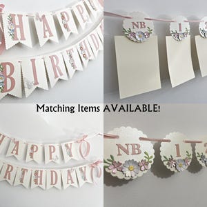 以下が含まれることがあります： ピンクと白の「Happy Birthday」の文字がピンクの文字で書かれたバナー。バナーは小さな白い花で飾られています。また、1、2、3、および「NB」の数字が書かれたマッチングカードもあります。