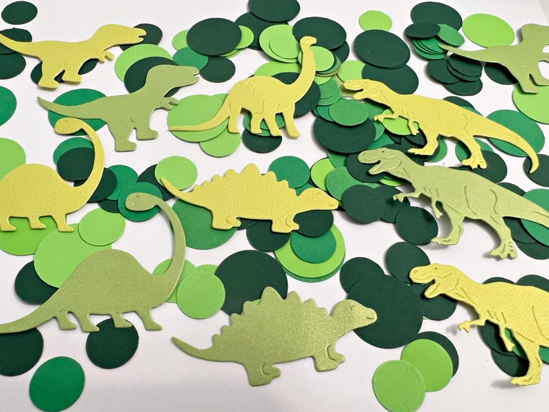 K&ouml;nnte beinhalten: Gr&uuml;ner und gelber Papier-Dinosaurier-Konfetti. Der Konfetti enth&auml;lt verschiedene Dinosaurierformen, darunter einen Stegosaurus, einen Brachiosaurus und einen Tyrannosaurus Rex.