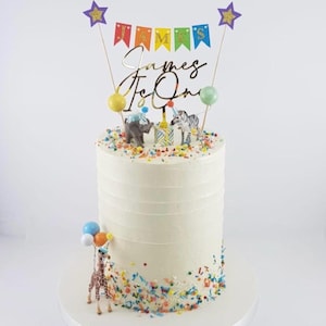 Cake topper that reads James Is One, a multi-colored pennant banner, two purple glitter stars