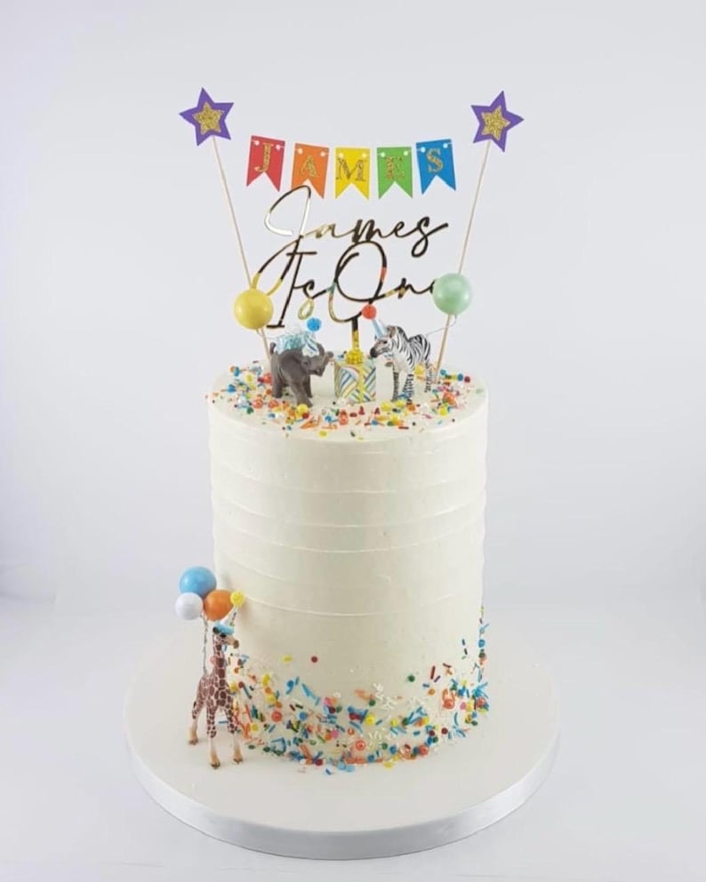 Cake topper that reads James Is One, a multi-colored pennant banner, two purple glitter stars