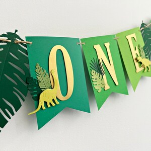 K&ouml;nnte beinhalten: Gr&uuml;nes und goldenes Banner mit dem Wort "ONE" in goldenen Buchstaben. Das Banner ist mit gr&uuml;nen Papierbl&auml;ttern und Dinosauriern verziert.
