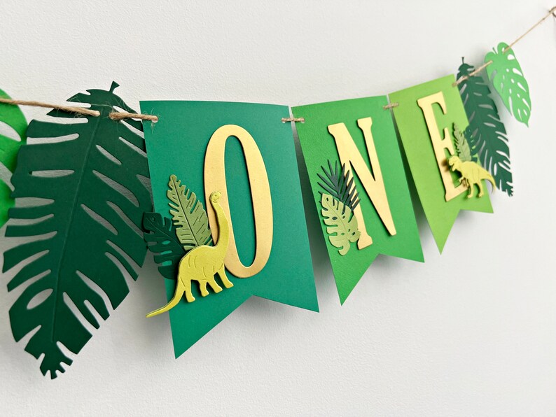 K&ouml;nnte beinhalten: Gr&uuml;nes und goldenes Banner mit dem Wort "ONE" in goldenen Buchstaben. Das Banner ist mit gr&uuml;nen Papierbl&auml;ttern und Dinosauriern verziert.