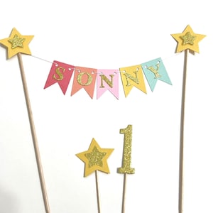 A cake topper set featuring a bunting banner with the name SONNY in gold glitter letters on multi-colored pennants, suspended between two wooden sticks topped with yellow stars, plus a gold glitter star and number 1 topper.
