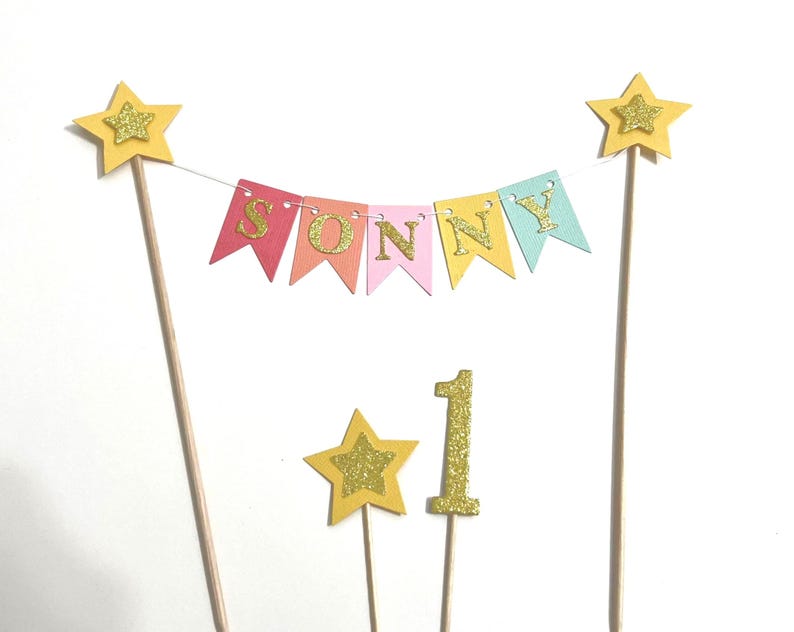 A cake topper set featuring a bunting banner with the name SONNY in gold glitter letters on multi-colored pennants, suspended between two wooden sticks topped with yellow stars, plus a gold glitter star and number 1 topper.