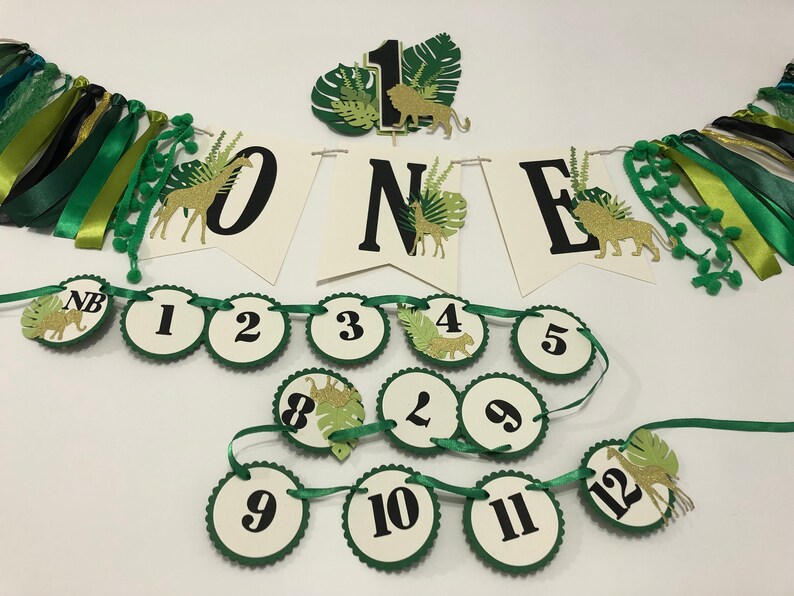 May include: Green and gold jungle-themed first birthday party decorations. The decorations include a banner with the word "ONE" in black letters, a cake topper with a lion, and a garland of green and gold circles with numbers from 1 to 12.