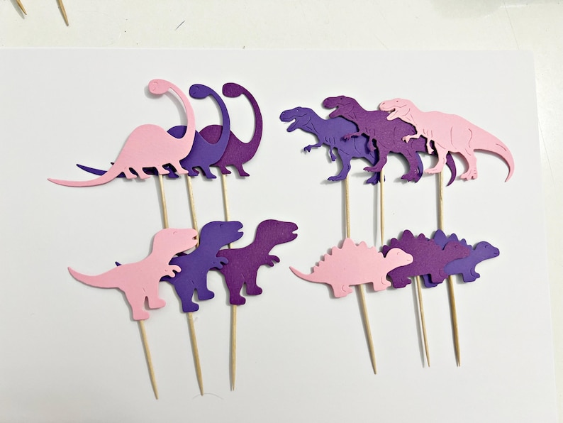 May include: Purple and pink dinosaur cake toppers. The dinosaurs are in various poses, including standing, walking, and running. They are made of cardstock and have wooden skewers attached to the back.
