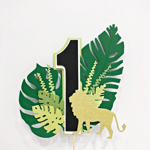 May include: Green and gold jungle-themed cake topper with a black number one, a gold glitter lion, and green leaves.