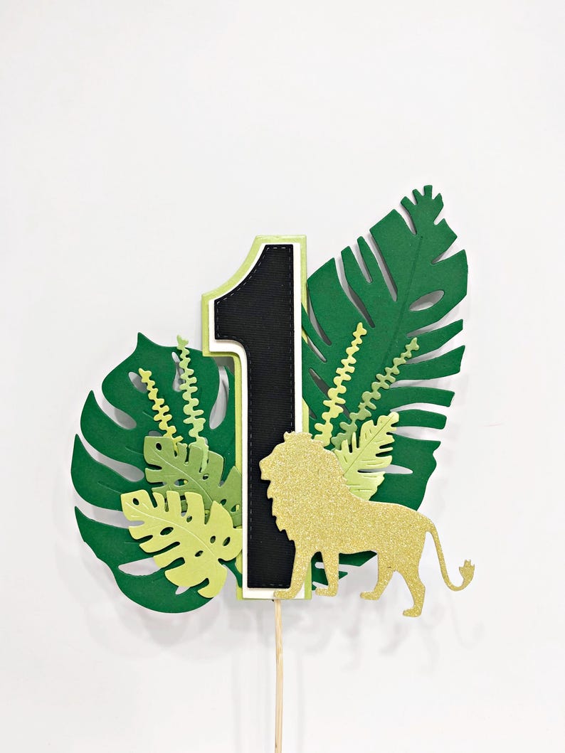 May include: Green and gold jungle-themed cake topper with a black number one, a gold glitter lion, and green leaves.