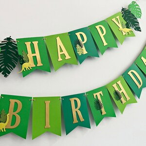 K&ouml;nnte beinhalten: Gr&uuml;n-goldene Girlande mit den Worten "Happy Birthday" in goldenen Buchstaben. Die Girlande ist mit gr&uuml;nen Bl&auml;ttern und Dinosauriersilhouetten verziert.