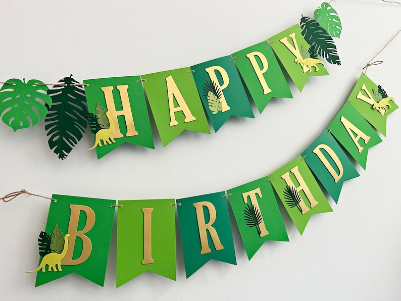 K&ouml;nnte beinhalten: Gr&uuml;n-goldene Girlande mit den Worten "Happy Birthday" in goldenen Buchstaben. Die Girlande ist mit gr&uuml;nen Bl&auml;ttern und Dinosauriersilhouetten verziert.