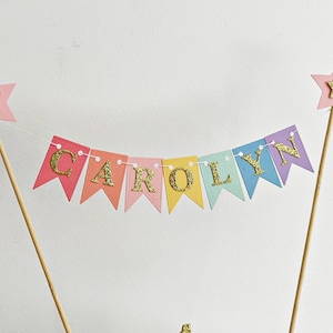 A cake topper banner featuring multi-colored pennant flags in pink, coral, yellow, mint, and lavender. Each flag displays a gold glitter letter spelling the name CAROLYN. The banner is suspended between two wooden sticks with pink stars.