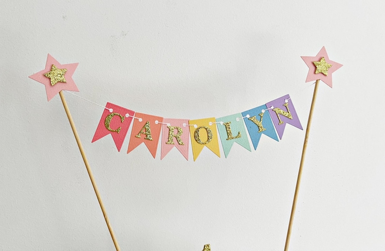 A cake topper banner featuring multi-colored pennant flags in pink, coral, yellow, mint, and lavender. Each flag displays a gold glitter letter spelling the name CAROLYN. The banner is suspended between two wooden sticks with pink stars.