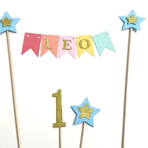 May include: A cake topper set featuring a bunting banner with pastel flags spelling LEO in gold glitter letters, two tall wooden sticks topped with light blue stars and gold glitter accents, a gold glitter number 1, and a small light blue star.