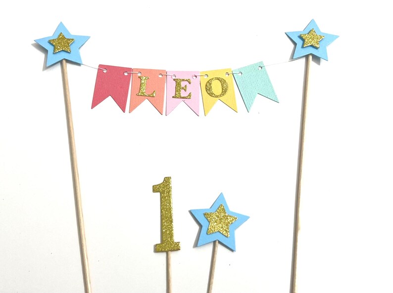 May include: A cake topper set featuring a bunting banner with pastel flags spelling LEO in gold glitter letters, two tall wooden sticks topped with light blue stars and gold glitter accents, a gold glitter number 1, and a small light blue star.