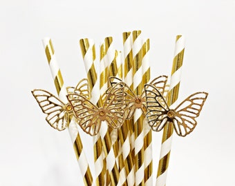 Könnte beinhalten: Ein Bündel Trinkhalme mit einem diagonalen Streifenmuster in Metallic-Gold und Weiß. Jeder Halm ist mit einem goldfarbenen, ausgestanzten Schmetterling auf der Vorderseite verziert, der filigrane Flügeldetails und einen kleinen zentralen Schmuckstein aufweist.