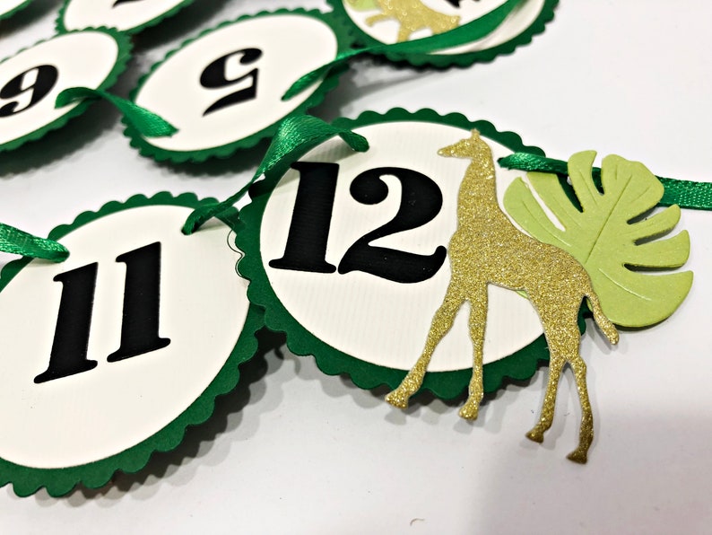 May include: Green and white paper circles with black numbers 9, 11, and 12. A gold glitter giraffe and a green leaf are attached to the circle with the number 12.