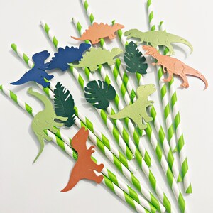 K&ouml;nnte beinhalten: Gr&uuml;n-wei&szlig; gestreifte Papierstrohhalme mit Dinosaurier- und Blattausschnitten. Die Dinosaurier sind in Gr&uuml;n-, Blau-, Orange- und Braunt&ouml;nen gehalten.