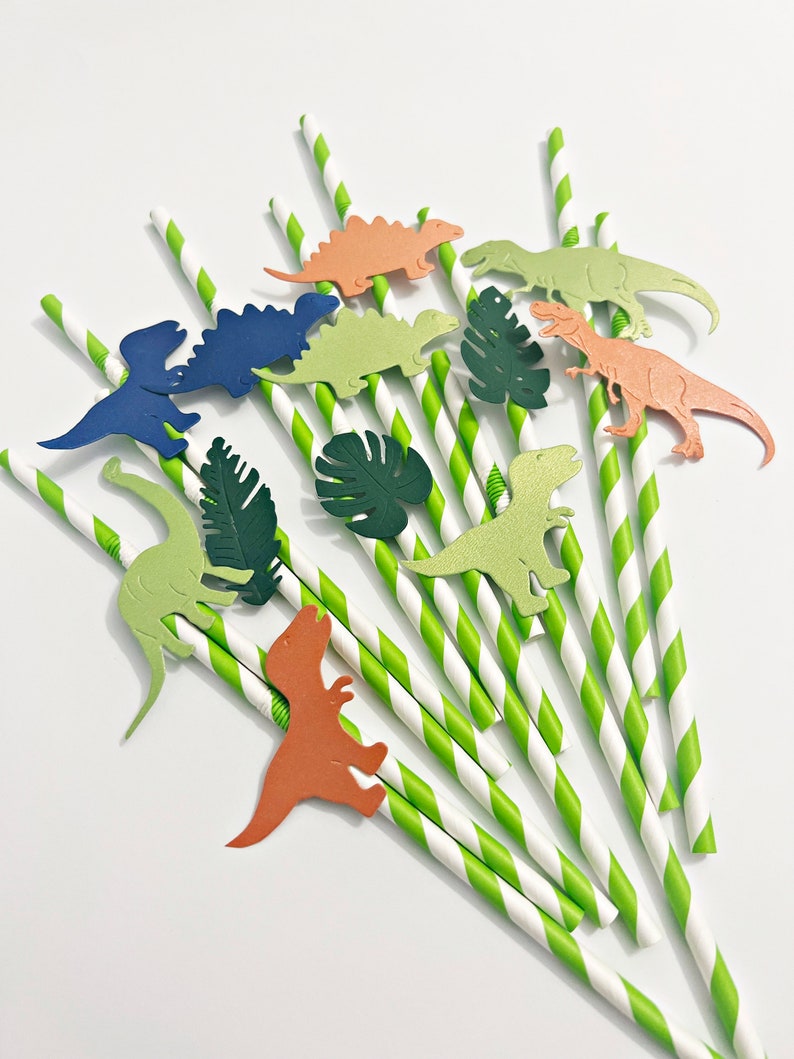 K&ouml;nnte beinhalten: Gr&uuml;n-wei&szlig; gestreifte Papierstrohhalme mit Dinosaurier- und Blattausschnitten. Die Dinosaurier sind in Gr&uuml;n-, Blau-, Orange- und Braunt&ouml;nen gehalten.