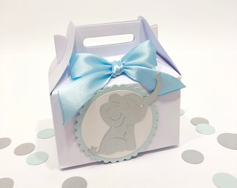 May include: A white favor box with a blue ribbon and a blue and gray elephant cutout. The box has a handle on top and is perfect for holding small gifts.