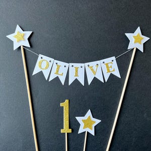 May include: A cake topper set featuring a white bunting banner with gold glitter letters spelling OLIVE, suspended between two wooden sticks topped with white and gold stars. Includes a separate gold glitter number 1 and a small star topper.