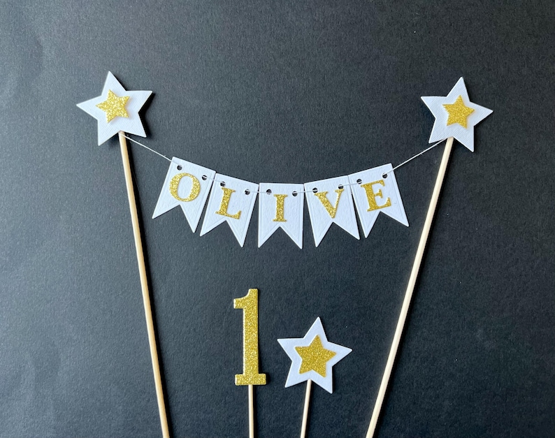May include: A cake topper set featuring a white bunting banner with gold glitter letters spelling OLIVE, suspended between two wooden sticks topped with white and gold stars. Includes a separate gold glitter number 1 and a small star topper.