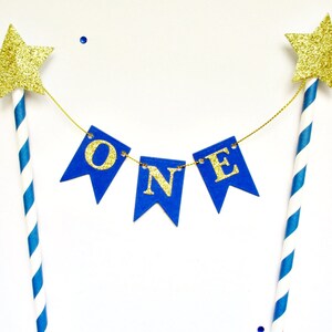 May include: A cake topper featuring two blue and white striped paper straws topped with gold glitter stars. A gold string connects the straws, holding three blue pennant flags that spell out the word ONE in gold glitter capital letters.
