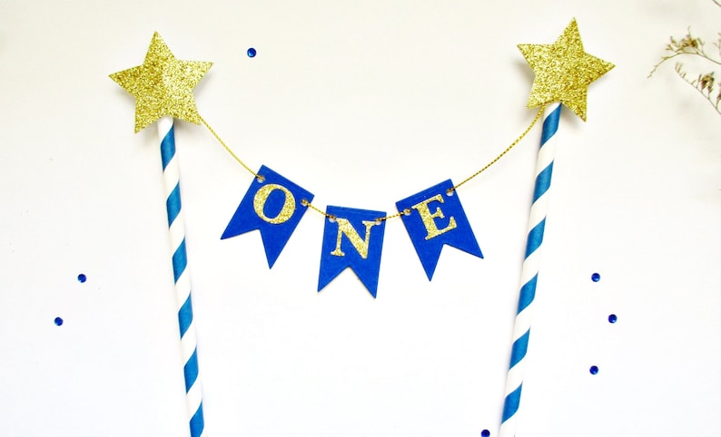 May include: A cake topper featuring two blue and white striped paper straws topped with gold glitter stars. A gold string connects the straws, holding three blue pennant flags that spell out the word ONE in gold glitter capital letters.