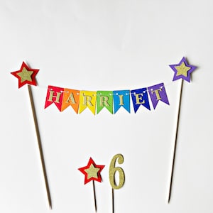 May include: A cake topper set featuring a rainbow-colored bunting banner with the name HARRIET in gold glitter letters, suspended between two wooden sticks topped with stars. Includes a separate gold glitter number 6 and a small star topper.
