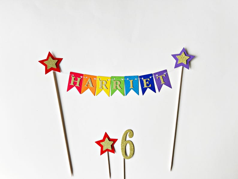 May include: A cake topper set featuring a rainbow-colored bunting banner with the name HARRIET in gold glitter letters, suspended between two wooden sticks topped with stars. Includes a separate gold glitter number 6 and a small star topper.