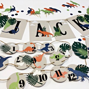 K&ouml;nnte beinhalten: Ein Dinosaurier-Party-Dekorationsset mit einem Banner, auf dem "JACK" steht, und einer Girlande aus 12 Kreisen mit Dinosauriern und Zahlen. Das Set enth&auml;lt auch gr&uuml;ne und braune Papierbl&auml;tter und einen blauen Dinosaurier-Topper.