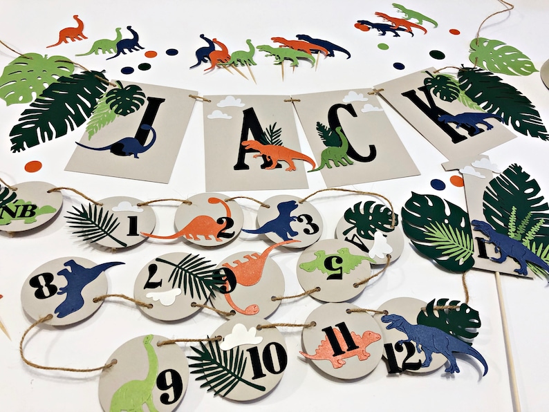 K&ouml;nnte beinhalten: Ein Dinosaurier-Party-Dekorationsset mit einem Banner, auf dem "JACK" steht, und einer Girlande aus 12 Kreisen mit Dinosauriern und Zahlen. Das Set enth&auml;lt auch gr&uuml;ne und braune Papierbl&auml;tter und einen blauen Dinosaurier-Topper.