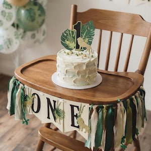 May include: A wooden high chair decorated with a banner that reads ONE, featuring green and cream fabric ribbons. A round cake with white frosting sits on the tray, topped with a cake topper showing a lion, palm leaves, and the number 1.