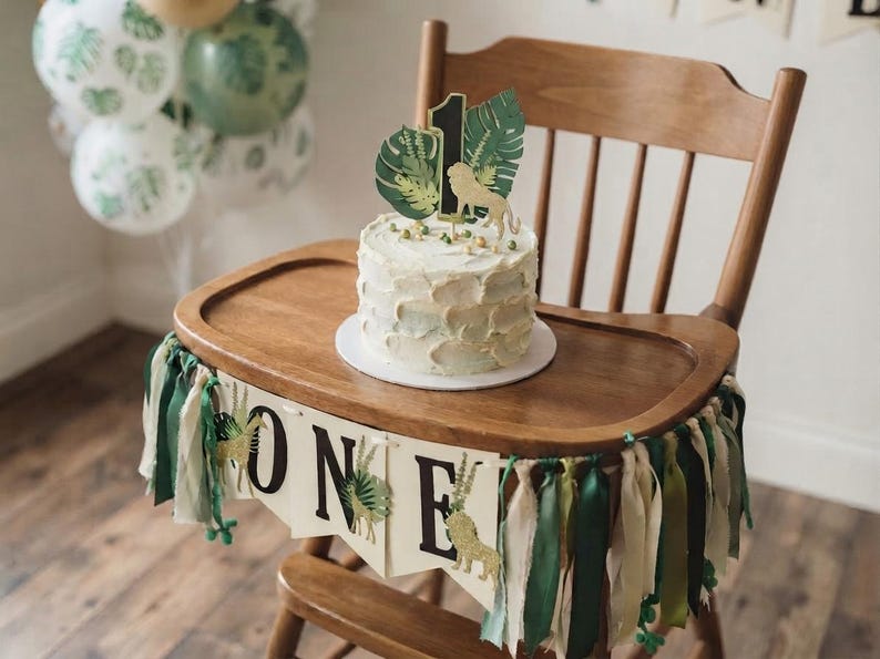 May include: A wooden high chair decorated with a banner that reads ONE, featuring green and cream fabric ribbons. A round cake with white frosting sits on the tray, topped with a cake topper showing a lion, palm leaves, and the number 1.