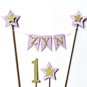 A cake topper set featuring two tall wooden sticks topped with light purple stars and gold glitter stars. A string connects the sticks, holding three light purple pennants with gold glitter letters spelling ZYN. A gold glitter number 1 and a small light purple star with a gold glitter star are also included.