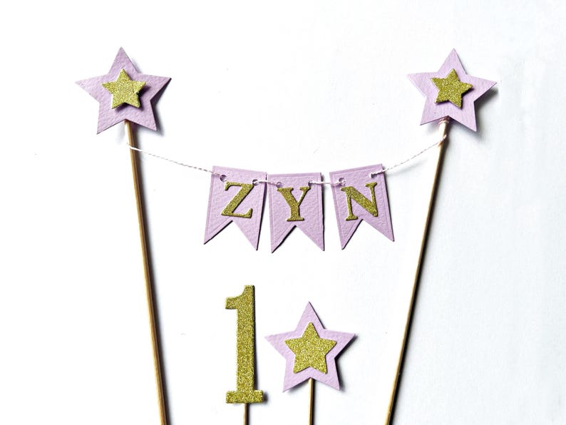 A cake topper set featuring two tall wooden sticks topped with light purple stars and gold glitter stars. A string connects the sticks, holding three light purple pennants with gold glitter letters spelling ZYN. A gold glitter number 1 and a small light purple star with a gold glitter star are also included.