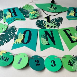 K&ouml;nnte beinhalten: Gr&uuml;ne und gelbe Dinosaurier-Geburtstagsdekorationen zum ersten Geburtstag. Das Banner lautet "Happy One" und enth&auml;lt gr&uuml;ne Bl&auml;tter und Dinosaurier. Es gibt auch gr&uuml;ne und gelbe Kreisdekorationen mit den Zahlen 1 bis 5.