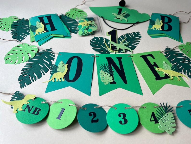 K&ouml;nnte beinhalten: Gr&uuml;ne und gelbe Dinosaurier-Geburtstagsdekorationen zum ersten Geburtstag. Das Banner lautet "Happy One" und enth&auml;lt gr&uuml;ne Bl&auml;tter und Dinosaurier. Es gibt auch gr&uuml;ne und gelbe Kreisdekorationen mit den Zahlen 1 bis 5.