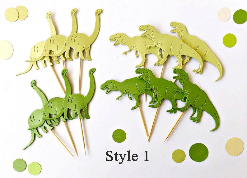 May include: A set of green and yellow dinosaur cake toppers. The toppers are made of paper and feature different types of dinosaurs, including a T-Rex and a Brontosaurus. The toppers are attached to wooden skewers. Style 1.