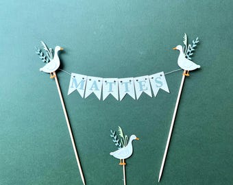 May include: A cake topper set featuring three white goose cutouts with orange beaks and feet, accented with light green foliage. Two geese are attached to wooden skewers, connected by a white string banner with the letters M A T T E S.