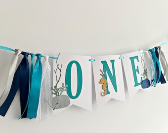 May include: A decorative banner featuring white pennants with the teal letters O, N, and E. The banner is accented with a gray whale, a yellow seahorse, and a blue whale cutout, all strung together with teal ribbon and navy, white, and lace fabric strips.